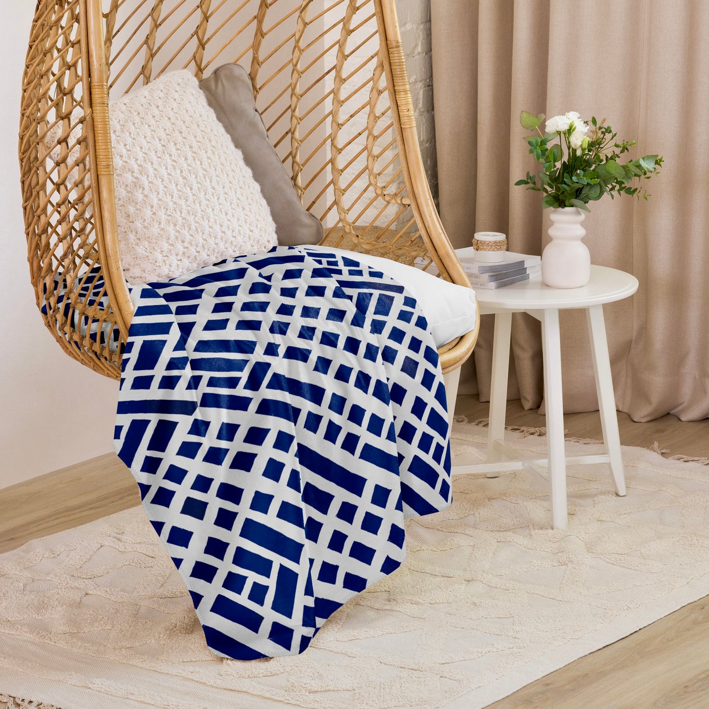 Tintin Studios' blue Felele Sherpa blanket draped elegantly over a wicker hanging chair, adding a touch of earthy warmth to a cosy, modern nook with a white side table and elegant curtains in the background.