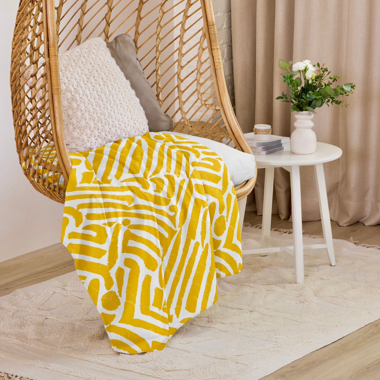 Vibrant yellow ChiChi Sherpa blanket from Tintin Studios draped over a stylish wicker hanging chair, adding a splash of colour and warmth to a cosy reading corner with a chic white side table and soft, neutral curtains in the background.