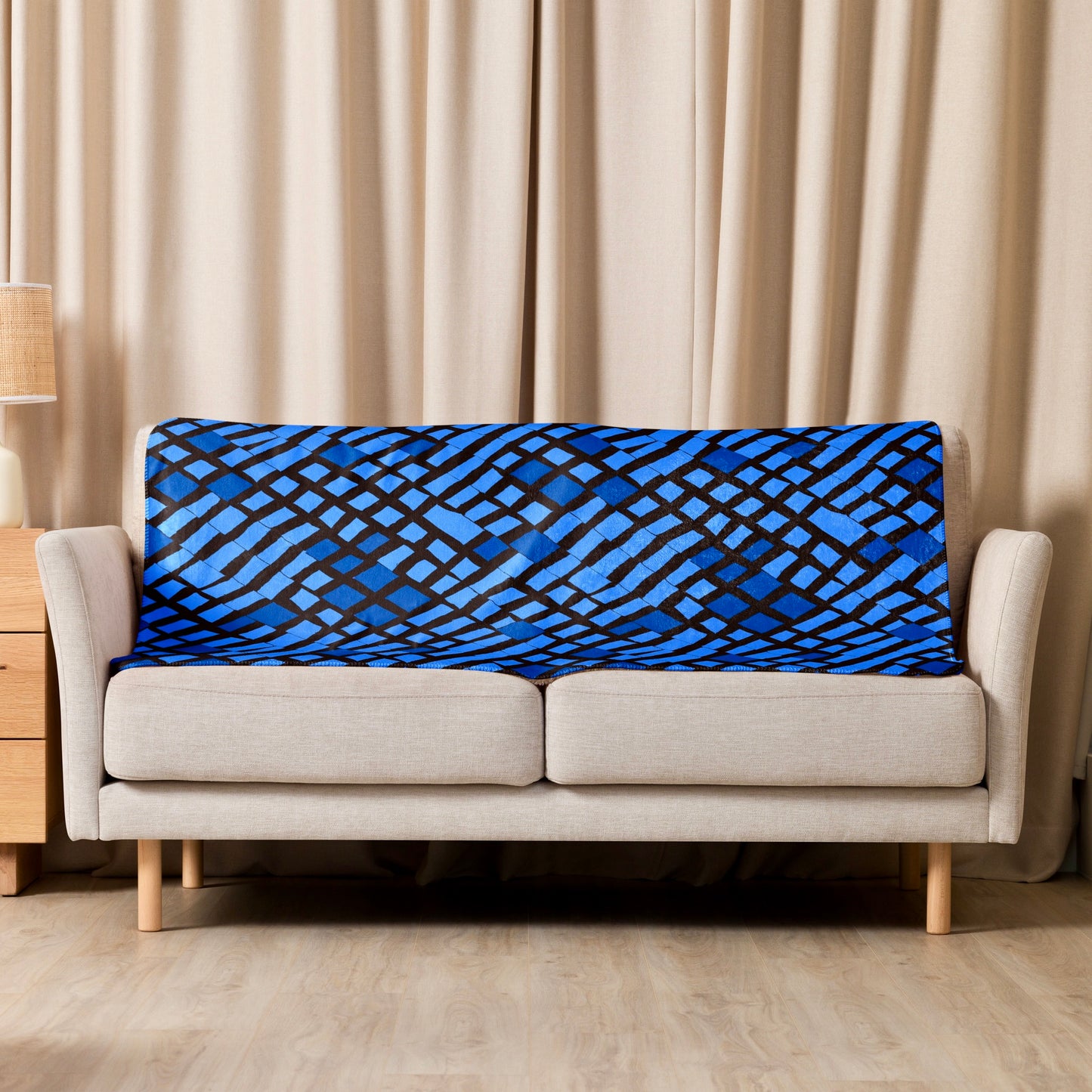 Blue Lagos Sherpa blanket from Tintin Studios with vibrant abstract designs thrown casually over a cream couch, providing a pop of colour and artistic flair to the living area.