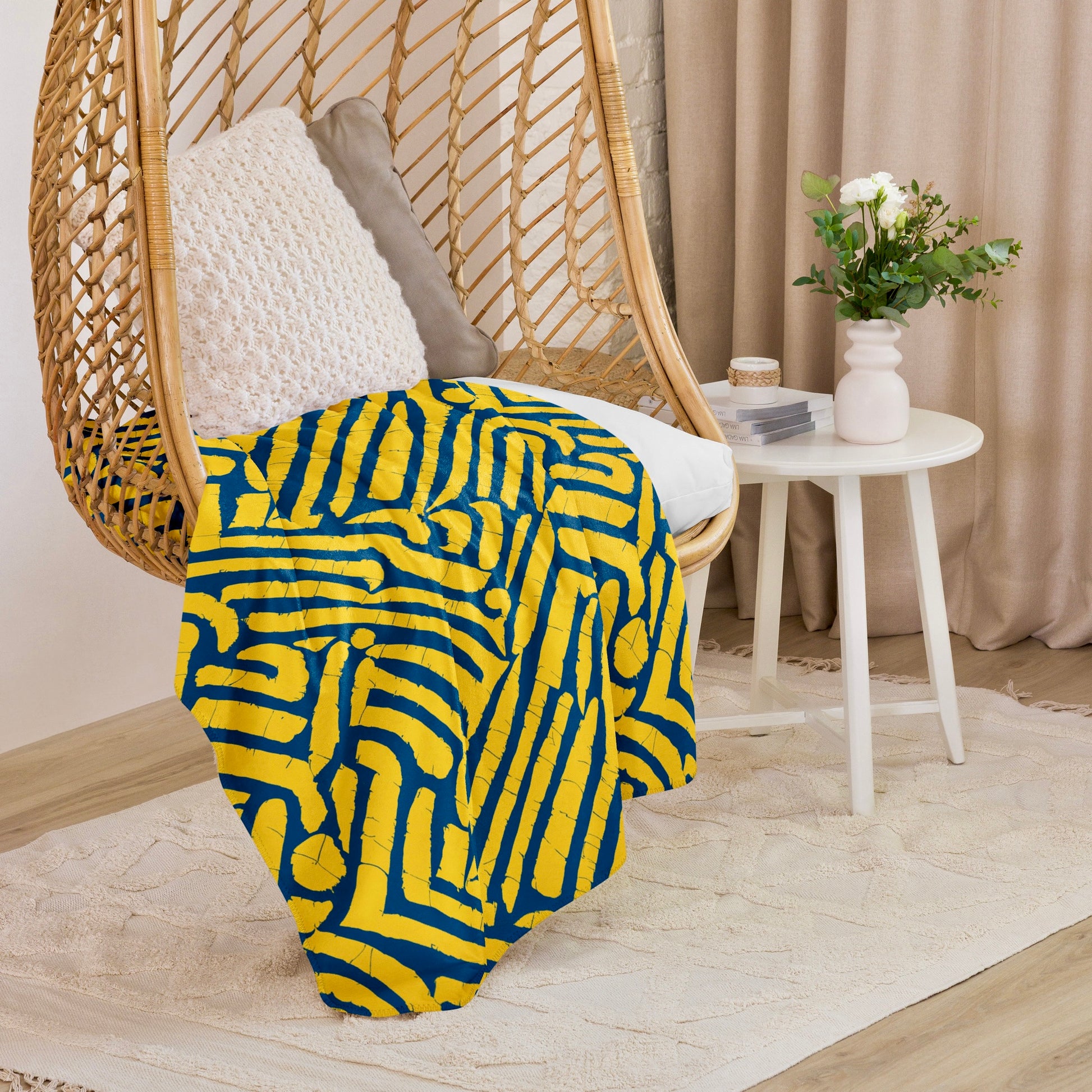 Vibrant blue ChiChi Sherpa blanket from Tintin Studios draped over a stylish wicker hanging chair, adding a splash of colour and warmth to a cosy reading corner with a chic white side table and soft, neutral curtains in the background.